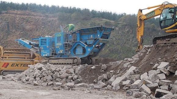 Recycling an airfield N03. The blue machine is a mobile crusher which is fed the large concrete by the yellow 360° degree excavator (JCB) on the right of shot. The crusher extracts much of the soil and then crushes the concrete into lumps around 2inch/50mil in size before feeding them out along the conveybelt on the ramp at the left end. From there the material either forms a cone for shifting elsewhere or as in this case it is fed directly into an Extec screener. Next stage 379759